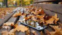 Smartphone noir posé sur un banc en bois, entouré de feuilles mortes d'automne aux teintes orangées et jaunes, avec un reflet du ciel sur l'écran du téléphone.