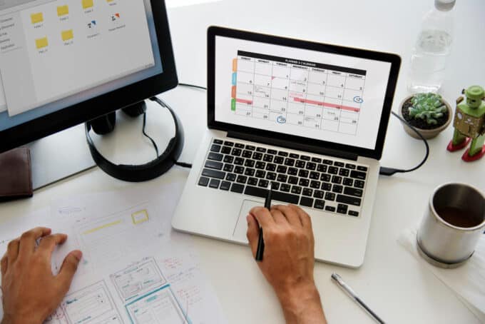 Personne travaillant à un bureau avec un ordinateur portable affichant un planning mensuel, tenant un stylo et une feuille avec des croquis, accompagné d'un écran secondaire, un casque audio, une tasse de café, une plante succulente et une bouteille d'eau.