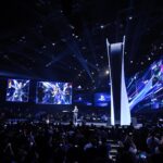 Conférence PlayStation avec une foule de spectateurs, un présentateur sur scène devant une immense reproduction stylisée de la PS5, des écrans géants affichant des visuels dynamiques et des logos PlayStation.