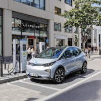 Voiture électrique sans permis en train de se recharger sur une borne 8K en ville, avec des passants et des bâtiments modernes en arrière-plan.