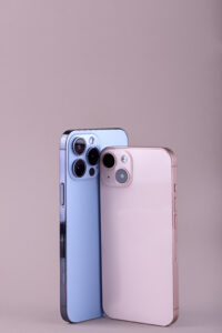 Deux smartphones modernes aux finitions brillantes, l’un bleu et l’autre rose, présentés verticalement sur fond uni rose pâle.